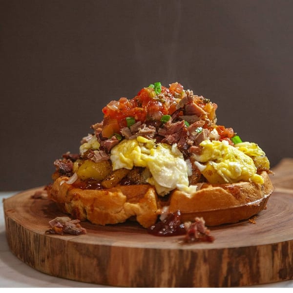 Savory waffle topped with scrambled eggs, potatoes, pulled pork, and fresh salsa on a wooden board.