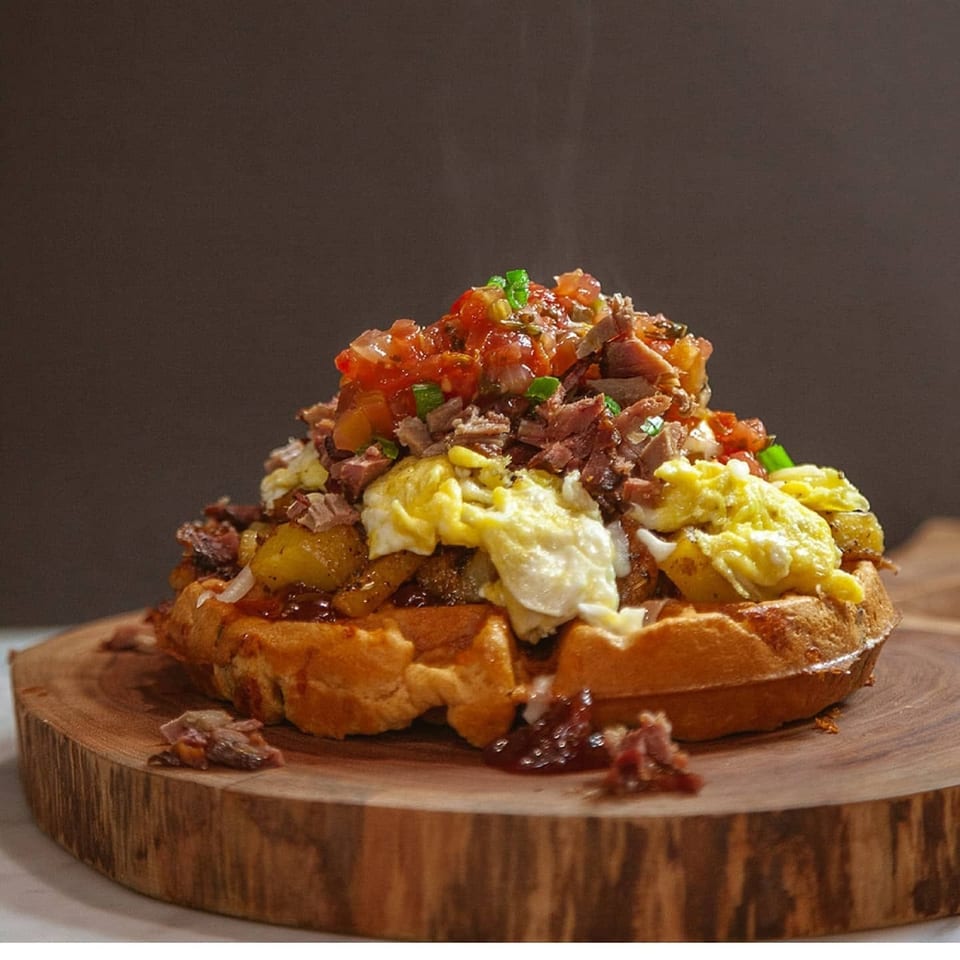 Savory waffle topped with scrambled eggs, potatoes, pulled pork, and fresh salsa on a wooden board.