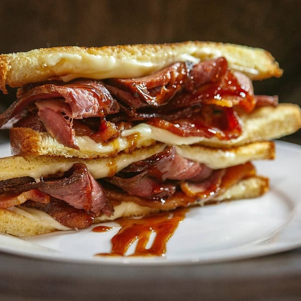 Stacked grilled cheese sandwich with melted cheese, pastrami, and a sweet glaze on a white plate.