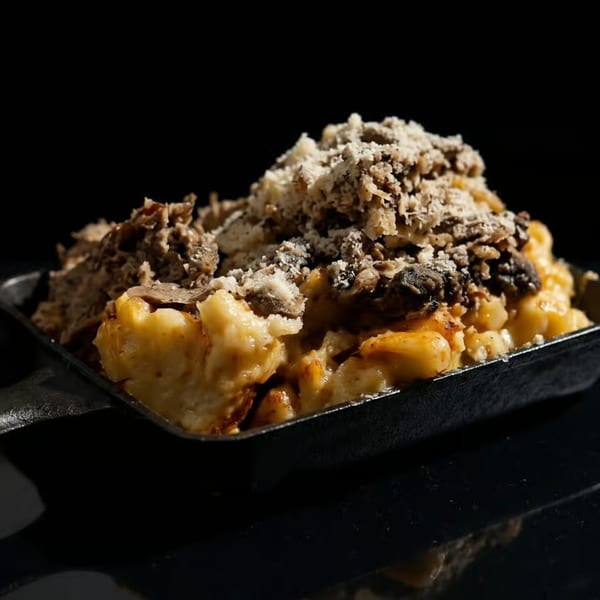 Close-up of mac and cheese topped with shredded meat, mushrooms, and grated cheese in a black pan.