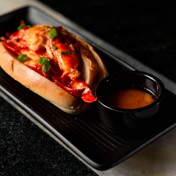 A lobster roll with green onions and a side of sauce on a black rectangular plate.