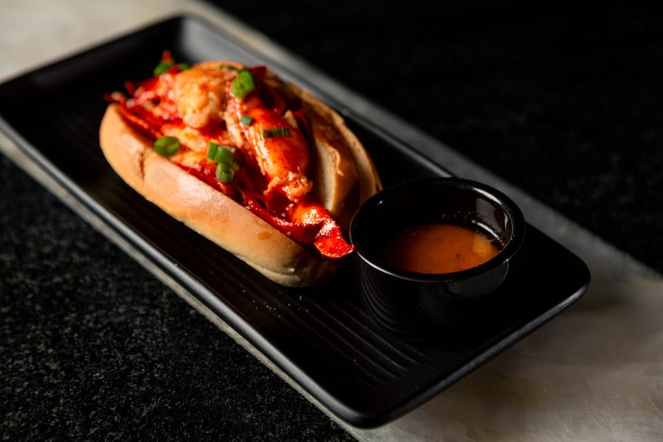 A lobster roll with green onions and a side of sauce on a black rectangular plate.