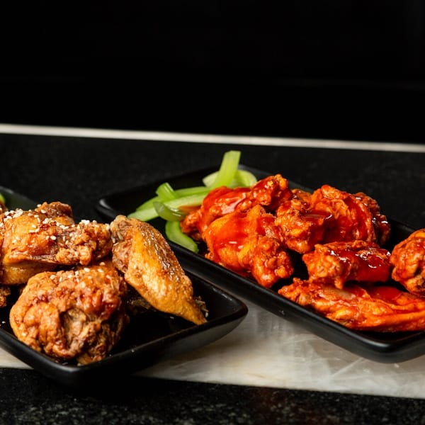 Two plates of chicken wings, one with sesame seeds and the other with red sauce, alongside celery sticks.