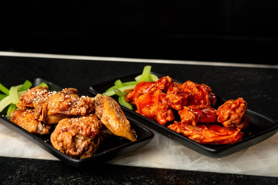 Two plates of chicken wings, one with sesame seeds and the other with red sauce, alongside celery sticks.