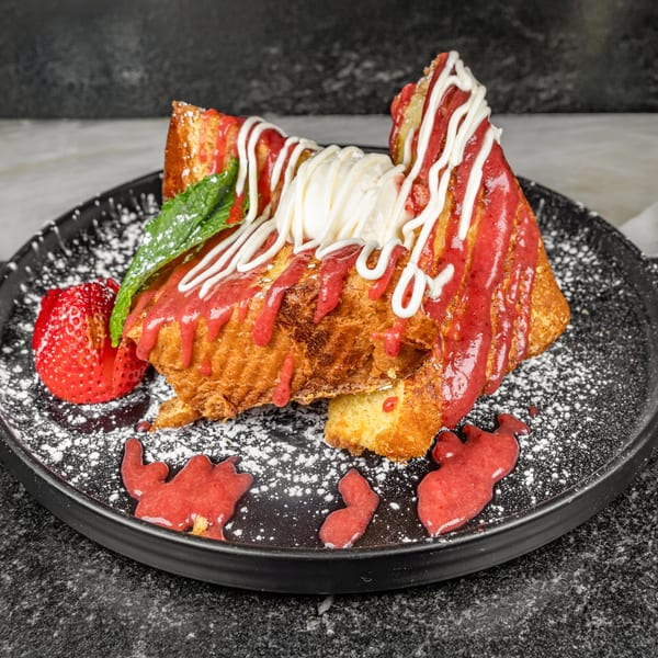 Thick-cut French toast with strawberry drizzle, whipped cream, powdered sugar, and a fresh strawberry.