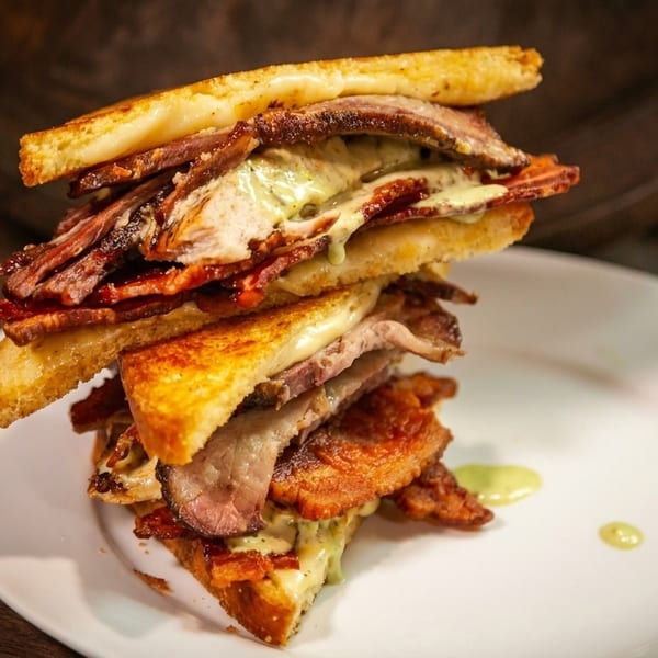 A towering sandwich with toasted bread, bacon, meat, and a creamy green sauce on a white plate.