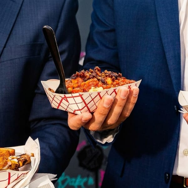 Two men in suits holding paper trays with chili and a grilled cheese sandwich.