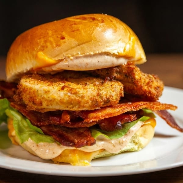Crispy chicken sandwich with bacon, lettuce, cheese, and sauce on a white plate.