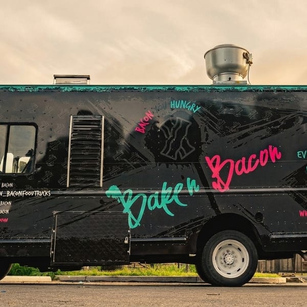 Black "Bake'n Bacon" food truck with pink and teal lettering parked outdoors under a cloudy sky.