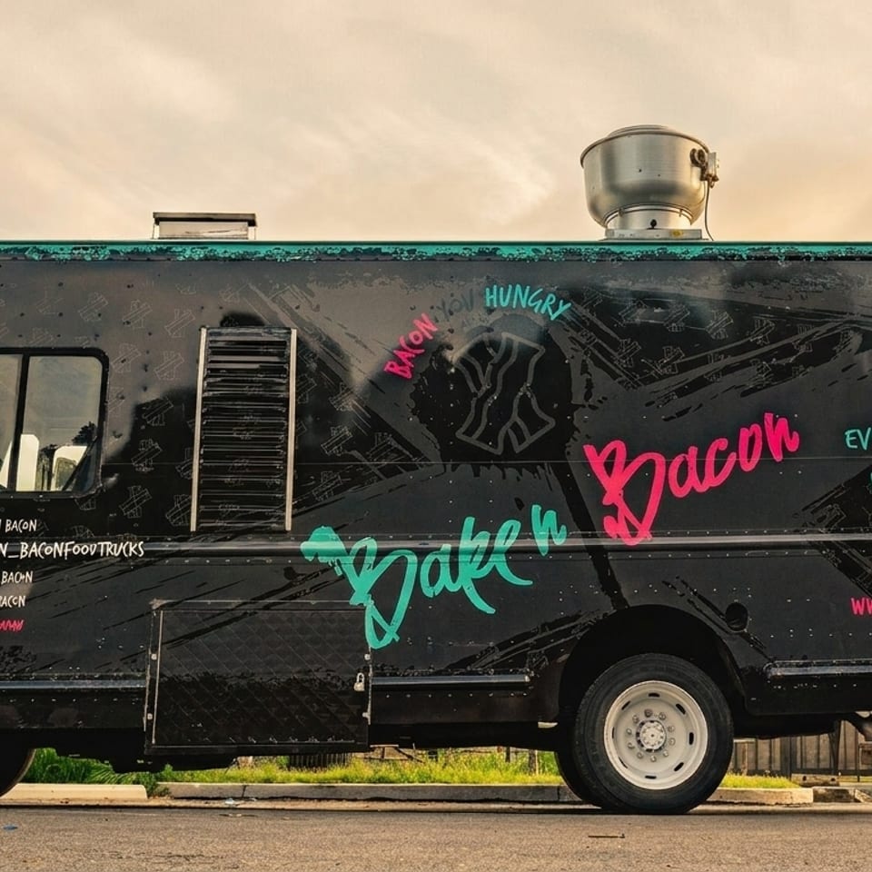 Black "Bake'n Bacon" food truck with pink and teal lettering parked outdoors under a cloudy sky.